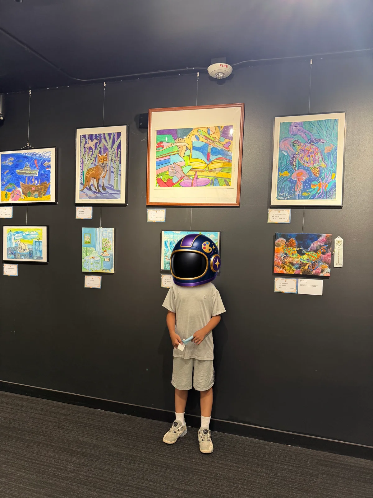 Eric standing beneath his displayed artwork Homework Break at the 2026 IAMA youth art exhibition, wearing an illustrated privacy helmet